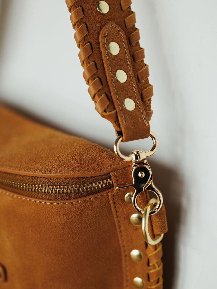 Close-up of The Yoakum by Savannah Sevens western life{&}style, a brown suede handbag with gold hardware, braided leather strap adorned with gold studs, decorative stitching, and a subtle snaffle bit accent. By Western and CO. 