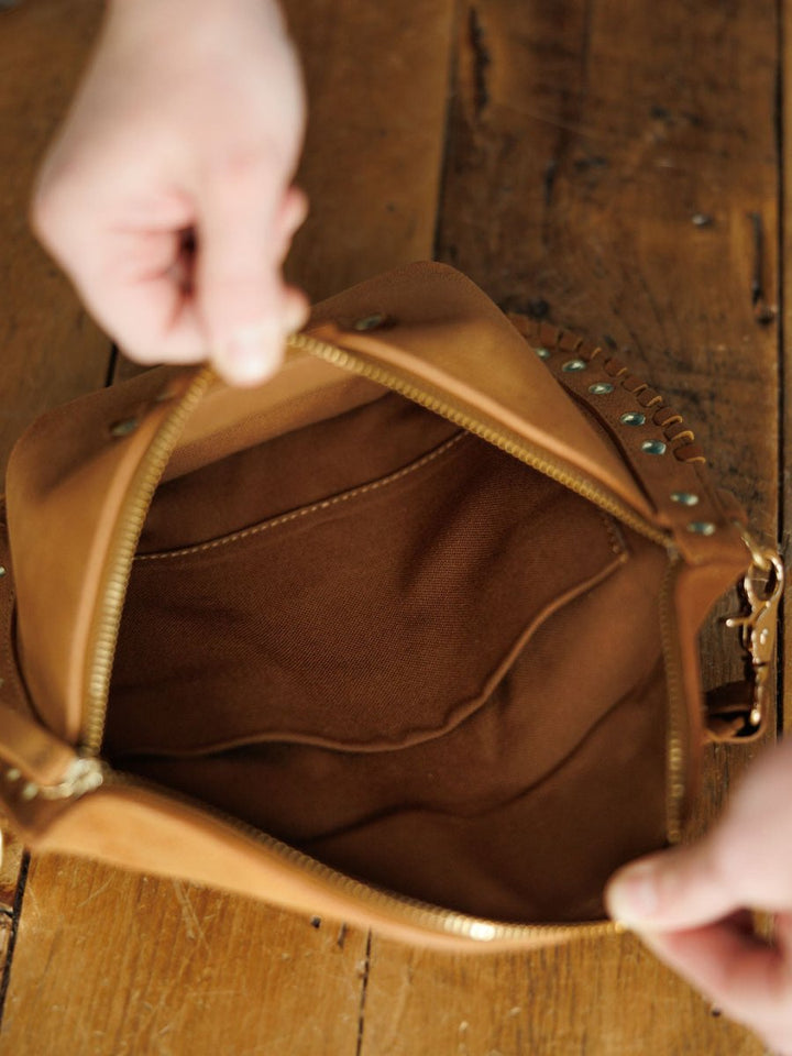 A person holds open The Yoakum leather bum bag by Savannah Sevens western life{&}style, displaying its suede interior and inner pocket on a wooden surface.