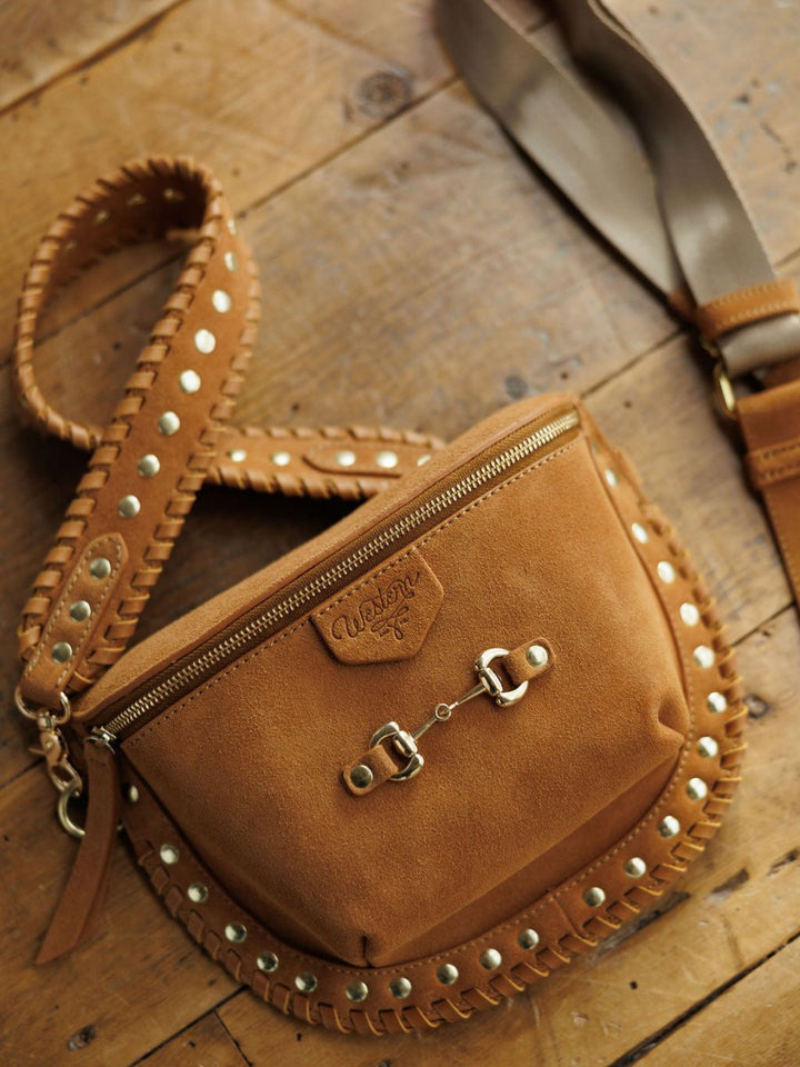 The Yoakum by Savannah Sevens western life{&}style is a brown leather crossbody bag with decorative stitching, metal studs, a zippered top, and branded patch, shown resting on a wooden surface.