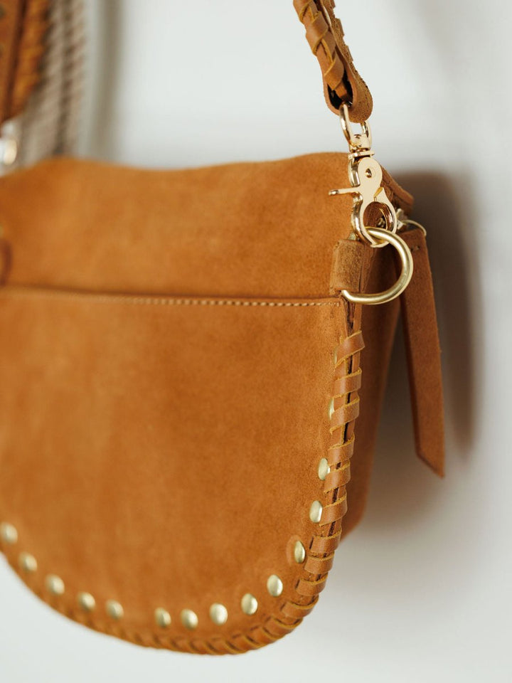 Close-up of The Yoakum, a tan suede handbag by Savannah Sevens western life{&}style, featuring gold studs along the edge, braided strap detailing, and snaffle bit hardware, set against a neutral background.