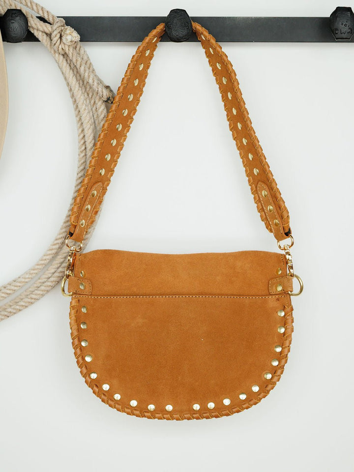 The Yoakum by Savannah Sevens western life{&}style—a tan suede handbag with a wide strap, gold studs, visible stitching, and snaffle bit accent—hangs on a black hook with a coiled rope against a white background. 