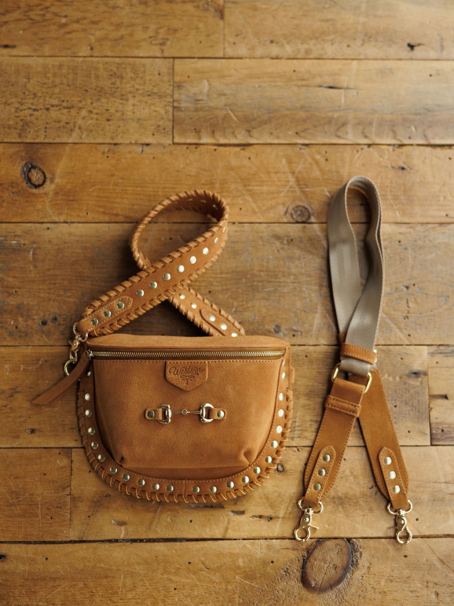 The Yoakum by Savannah Sevens western life{&}style is a tan leather crossbody with gold studs, detailed stitching, a snaffle bit detail, gold buckles, and two detachable straps—one brown studded leather, one beige fabric with leather ends.