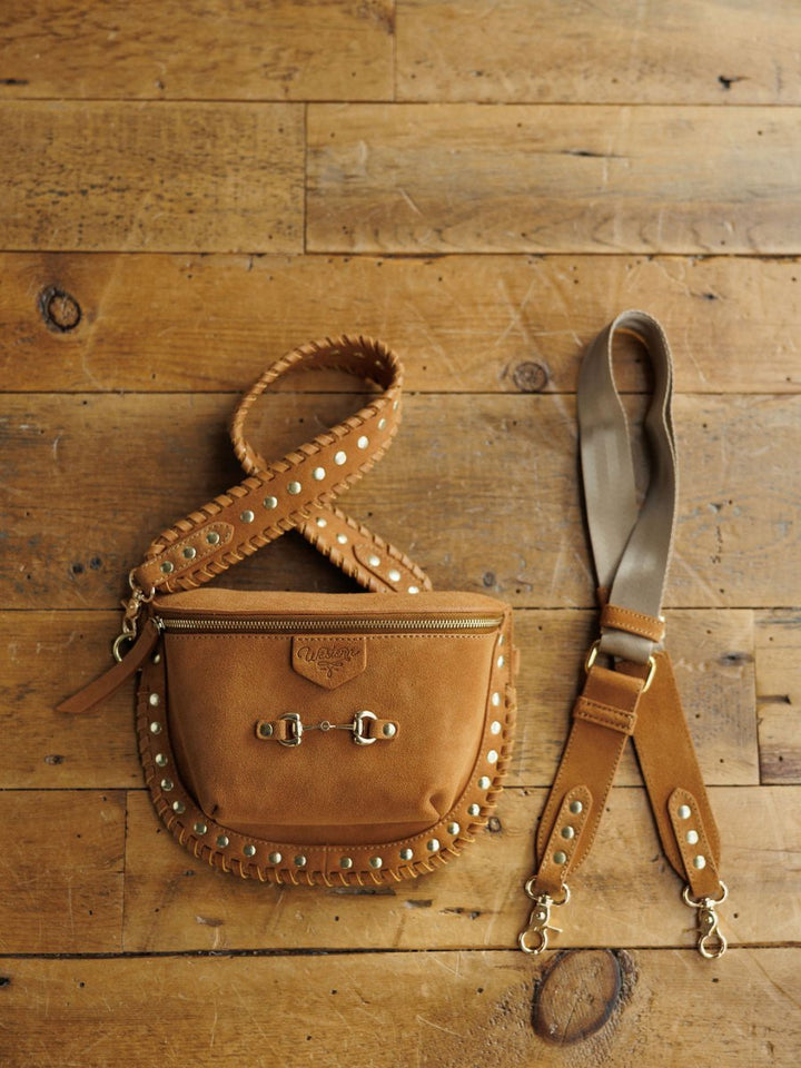 The Yoakum by Savannah Sevens western life{&}style is a tan leather crossbody with gold studs, detailed stitching, a snaffle bit detail, gold buckles, and two detachable straps—one brown studded leather, one beige fabric with leather ends.