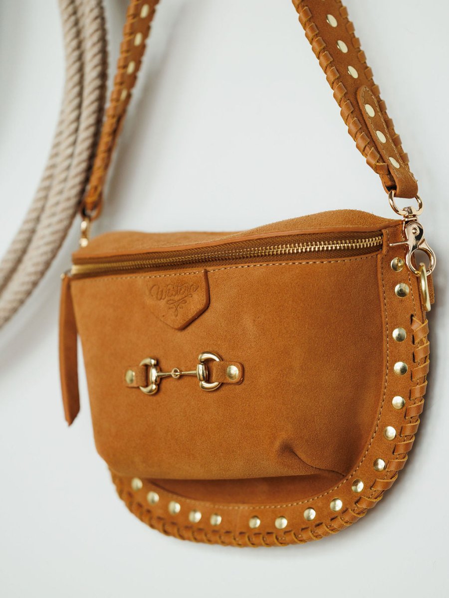 The Yoakum by Savannah Sevens western life{&}style is a brown suede crossbody bag with gold studs, decorative hardware, a snaffle bit accent, zipper closure, and stitched edges shown against a white background.
