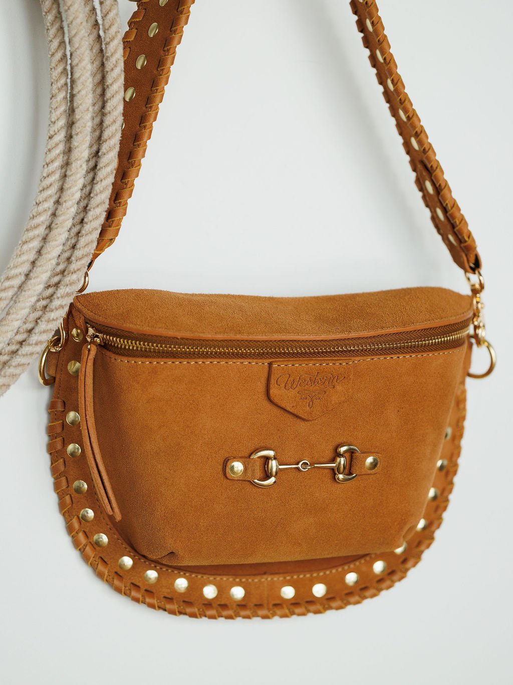 The Yoakum crossbody bag by Savannah Sevens western life{&}style, with tan suede, gold studs, snaffle bit detail, and stitched edges, hangs on a white wall. A looped rope in the background adds a Western vibe to the scene.