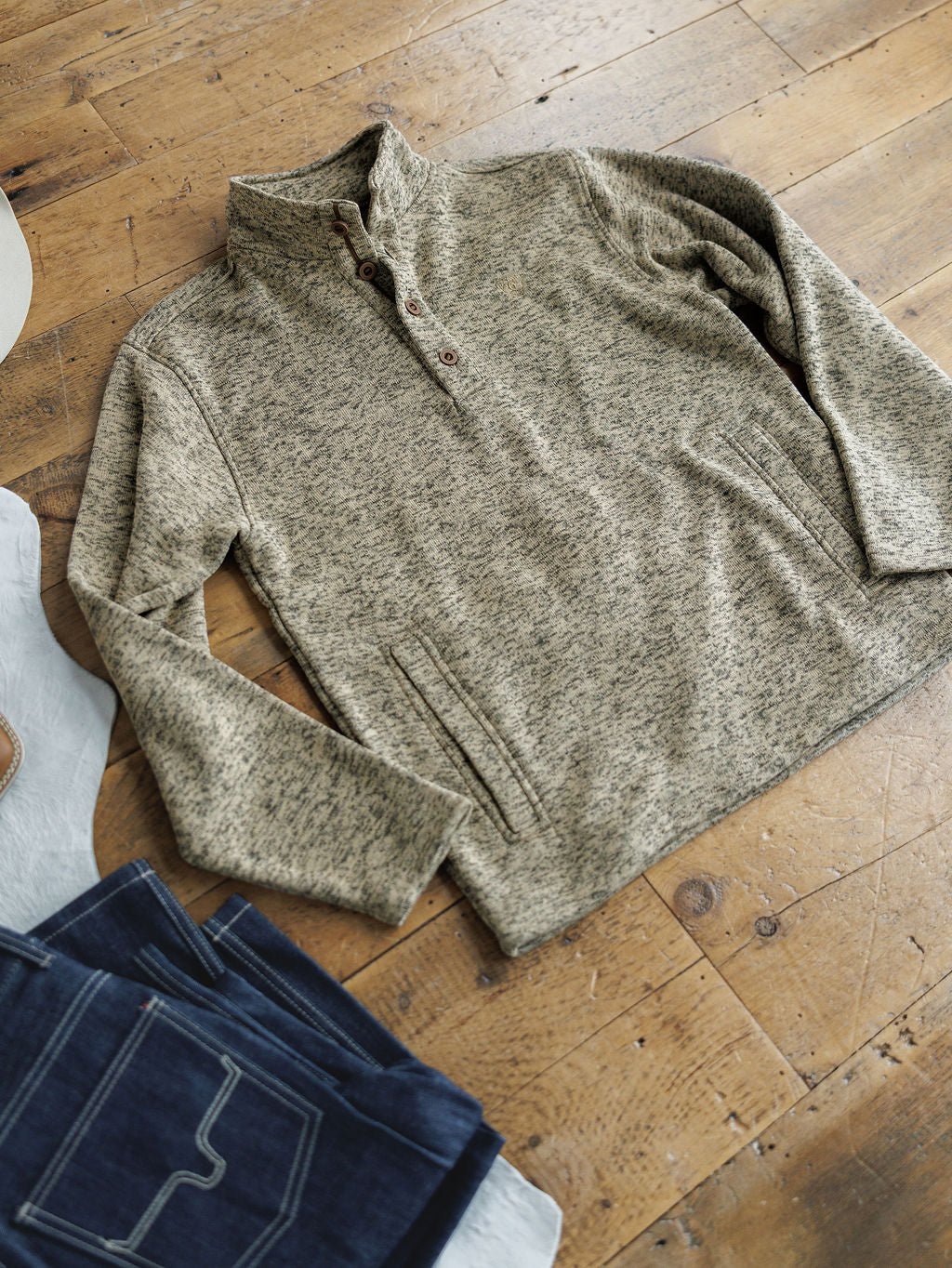 A Thompson Ariat Men's Pullover by Savannah Sevens western life{&}style in fleece-lined heathered beige with buttons is displayed on a wooden floor, next to folded blue jeans and a white cowboy hat.
