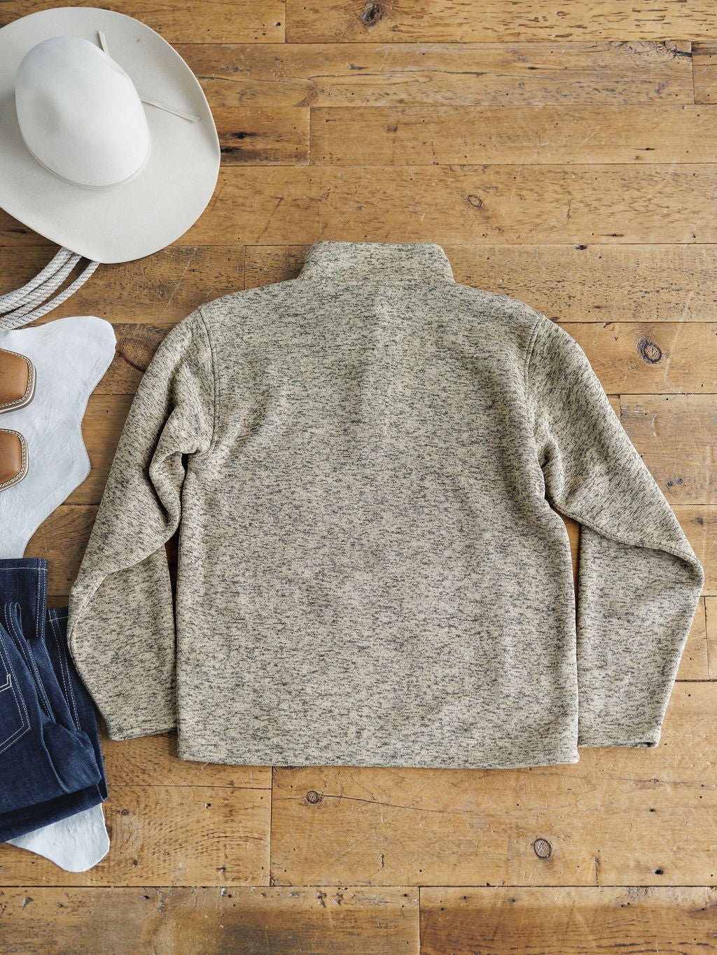 A Thompson Ariat Men's Pullover by Savannah Sevens western life{&}style in heathered gray is laid flat on a wooden floor, with a white cowboy hat, white boots with tan accents, and blue jeans. The fleece lining provides comfort and moisture-wicking performance.