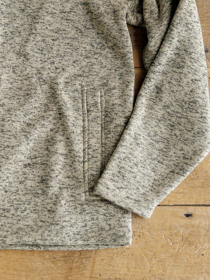 Close-up of a Thompson Ariat Men's Pullover by Savannah Sevens western life{&}style—a fleece-lined, long-sleeve light gray and black speckled sweater with side pocket, laid flat on wood. Only part of the body and one sleeve are shown.