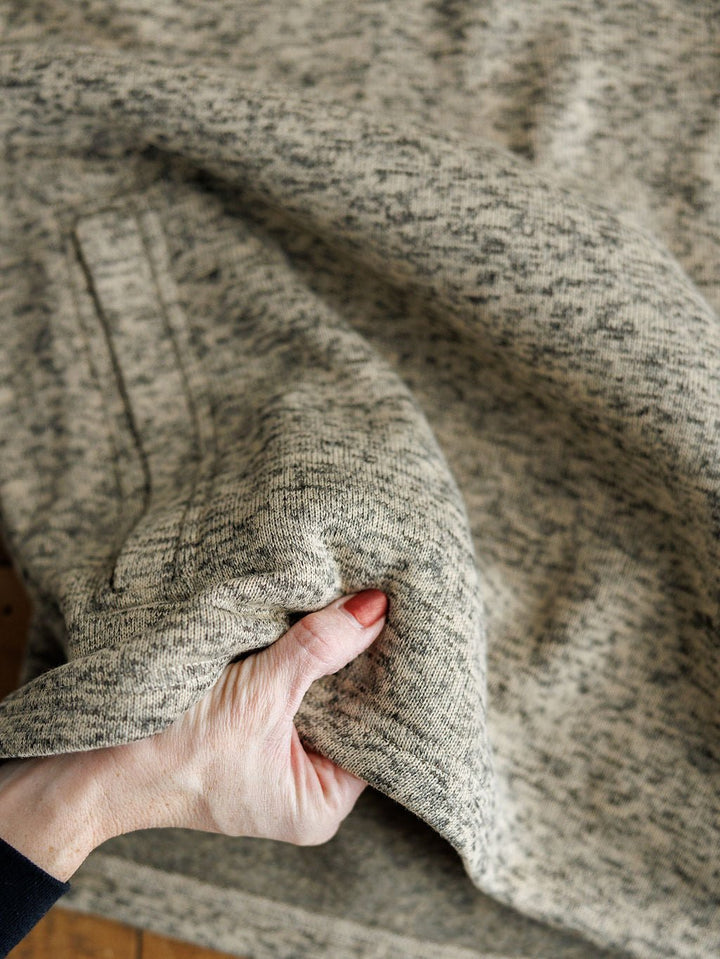 A hand lifts the soft, fleece-lined heathered beige and gray knit fabric of the Thompson Ariat Men's Pullover by Savannah Sevens western life{&}style, highlighting its thickness, moisture-wicking quality, and texture.