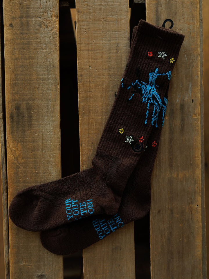 The Tie One On Crew Socks by Savannah Sevens western life{&}style are dark brown with blue embroidered text, a rider on horseback, and colored stars. Made from a comfy odor-fighting hemp blend, they're shown on a rustic wooden surface.