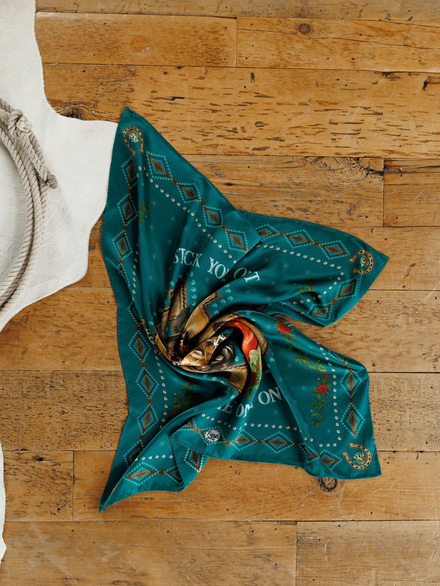 The Tie One On Neck Scarf by Savannah Sevens western life{&}style, in teal silk with ornate patterns and text, is artfully displayed in a spiral on wood flooring beside a white rug and rope, capturing western show horse elegance.
