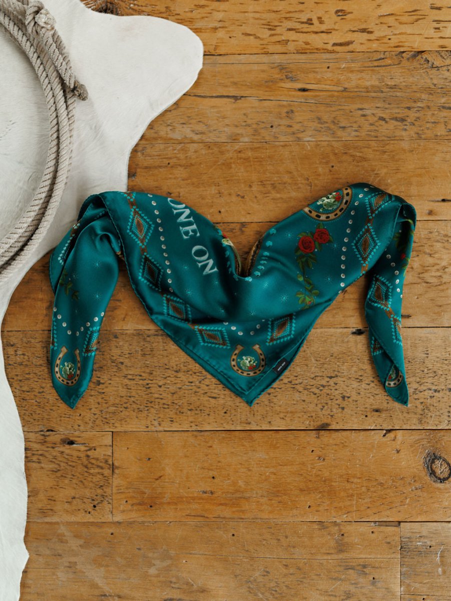 The Tie One On Neck Scarf by Savannah Sevens western life{&}style, a teal silk scarf with "RIDE ON" embroidery, is heart-folded on wood next to a cowhide rug—an ideal accent for any western show horse look.
