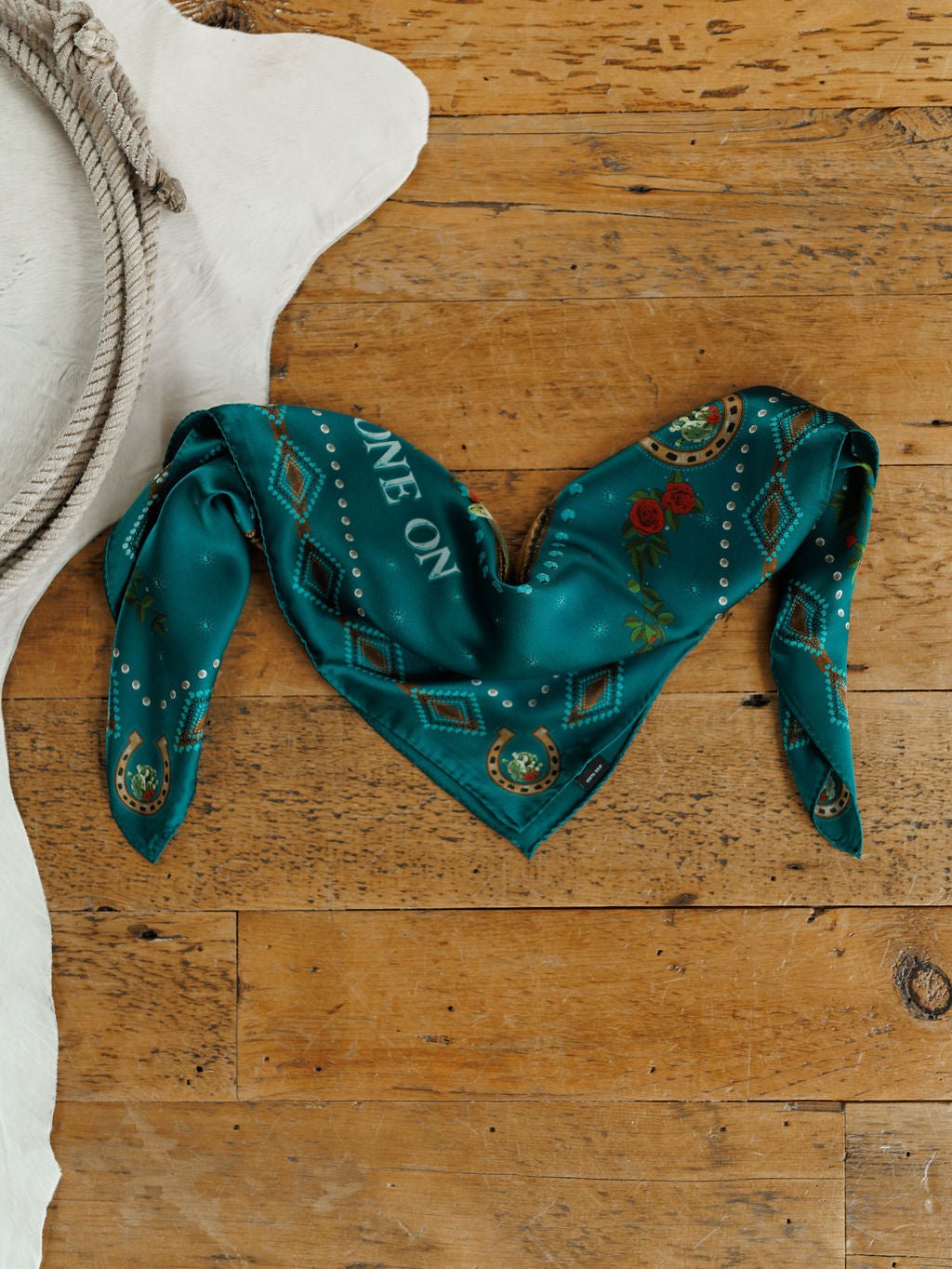 The Tie One On Neck Scarf by Savannah Sevens western life{&}style, a teal silk scarf with "RIDE ON" embroidery, is heart-folded on wood next to a cowhide rug—an ideal accent for any western show horse look.