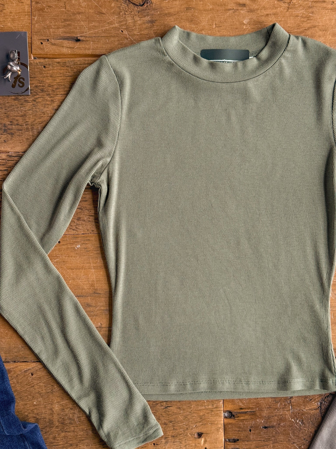 The Tifton Silver Sage Ribbed Basic Top by Savannah Sevens western life{&}style, a long-sleeve ribbed style in a subtle silver sage tone, is displayed lying flat on a wooden surface. 