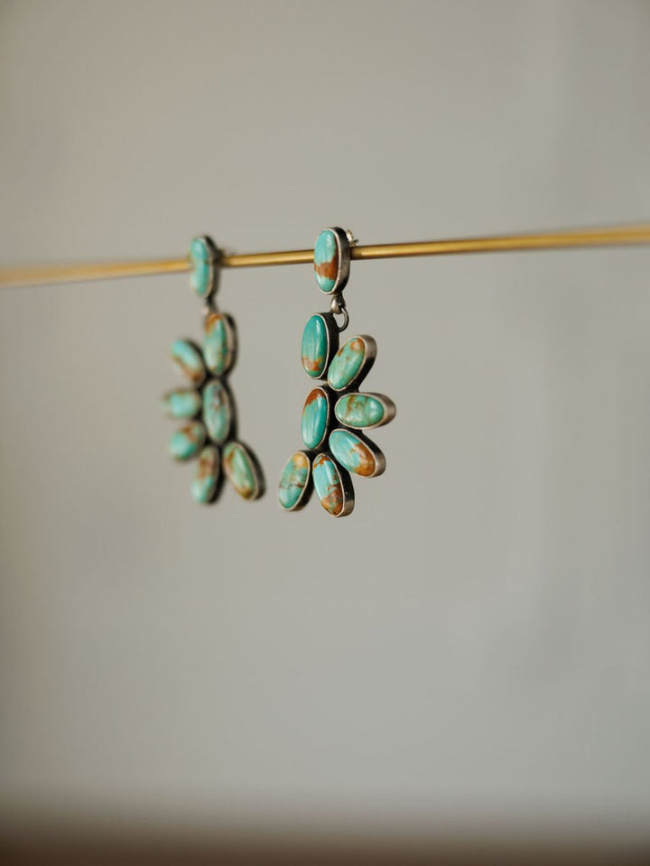 The Tijeras Earrings by Savannah Sevens western life{&}style feature Kingman turquoise oval stones in a floral pattern, suspended from thin gold rods and showcased against a neutral background.
