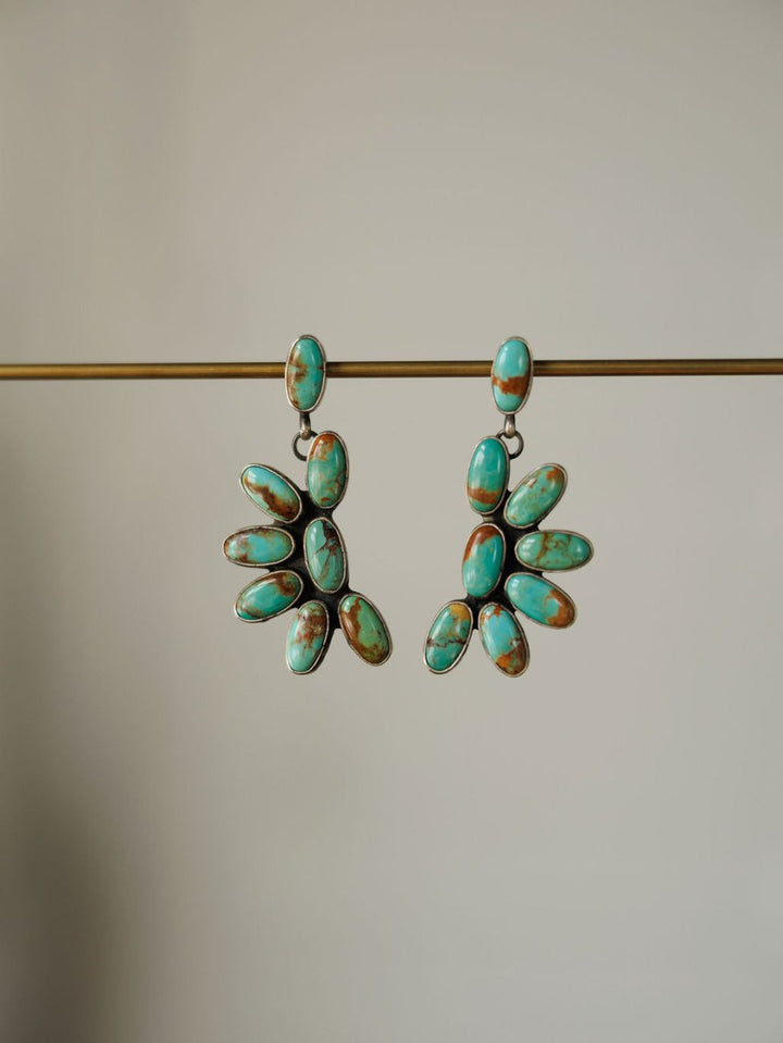 The Tijeras Earrings by Savannah Sevens western life{&}style feature dangling sterling silver with clusters of oval Kingman turquoise in a fan pattern, showcased on a plain beige background.