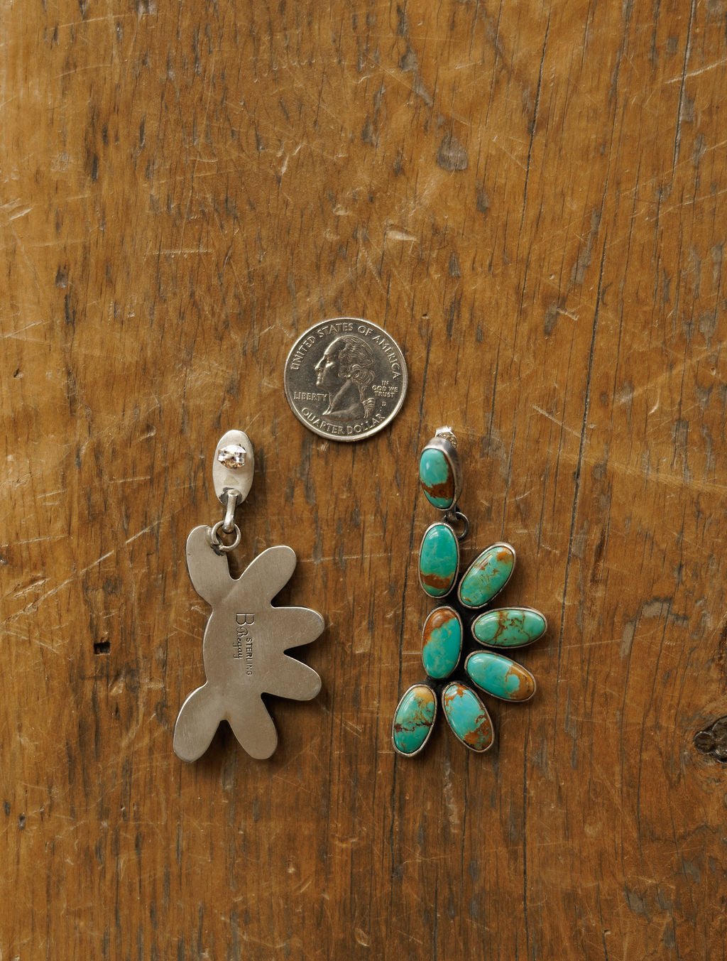 A US quarter is set between two Tijeras Earrings by Savannah Sevens western life{&}style on wood; the left earring shows a silver abstract back, while the right displays Kingman turquoise in a floral silver setting.