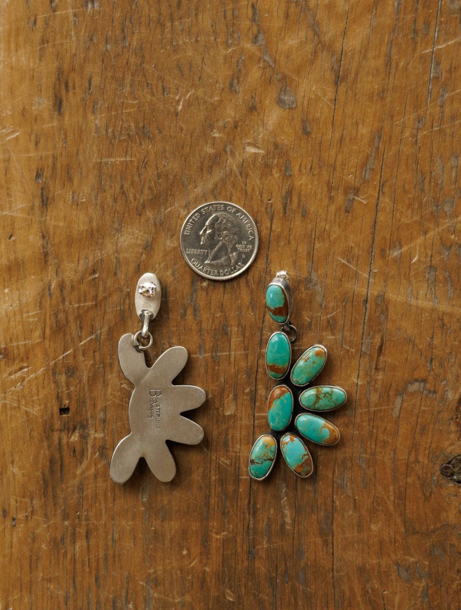 A US quarter is set between two Tijeras Earrings by Savannah Sevens western life{&}style on wood; the left earring shows a silver abstract back, while the right displays Kingman turquoise in a floral silver setting.