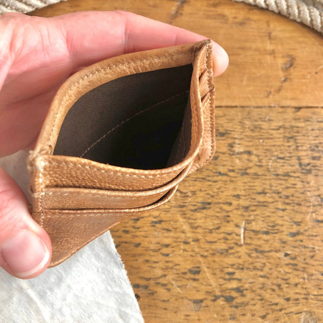 A hand holds the Tilden Wallet by Savannah Sevens western life{&}style—an empty rustic leather wallet with card slots—over a wooden surface.