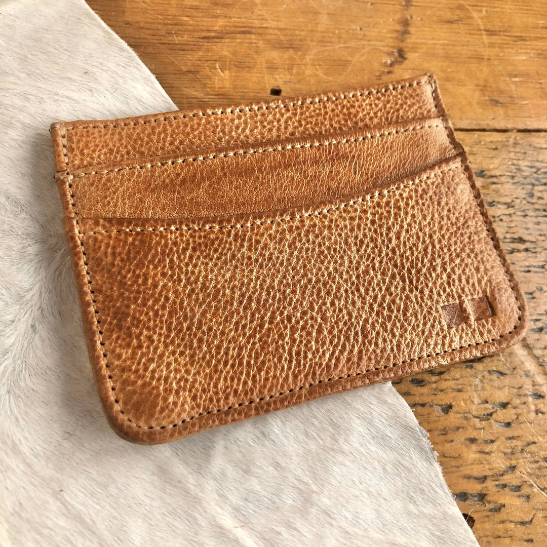 The Tilden Wallet by Savannah Sevens western life{&}style, a brown genuine leather card wallet with two slots, sits on a white and brown cowhide surface atop a wooden table.