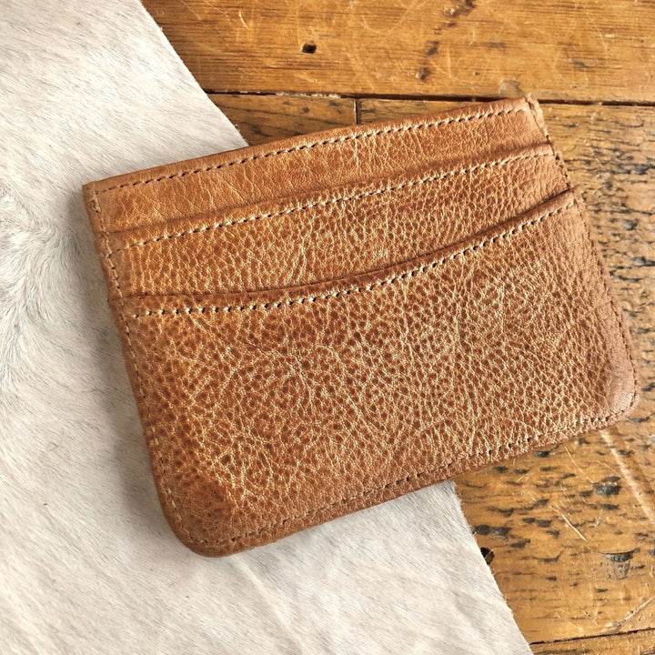 The Tilden Wallet by Savannah Sevens western life{&}style, a light brown genuine leather cardholder with visible stitching, rests on a white surface beside a rustic wooden table.