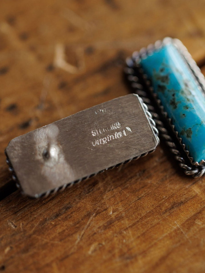 A close-up of Toano Earrings by Savannah Sevens western life{&}style and Navajo handcrafted earrings with a blue-green stone, both resting on a wooden surface.