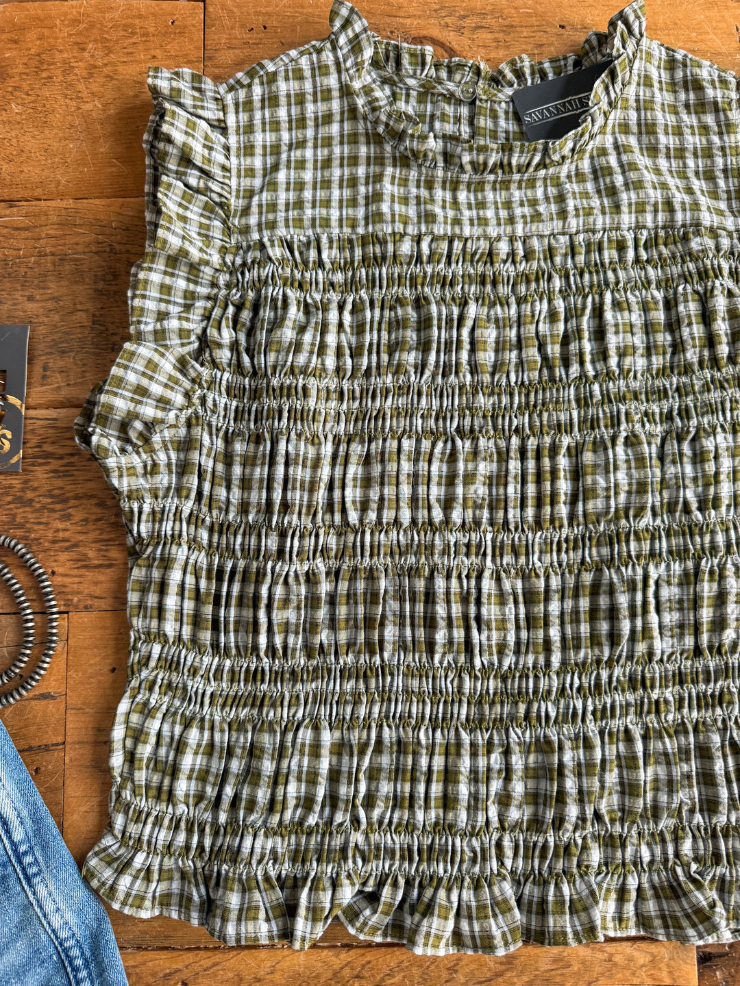 The Tolar Olive Plaid Top by Savannah Sevens western life{&}style, featuring a green and white plaid pattern with a sleeveless design, ruffle neckline, and smocked detailing, is laid flat on a wooden surface. Edges of the image show part of a coiled hair tie and blue denim fabric.