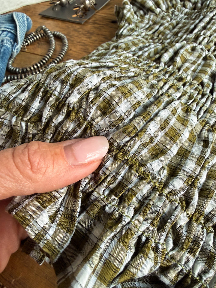 A hand with manicured nails holds a section of green and white plaid, ruched fabric from the Tolar Olive Plaid Top by Savannah Sevens western life{&}style, highlighting the textured, elastic shirred design. Jewelry and other items are softly blurred in the background on a wooden surface.