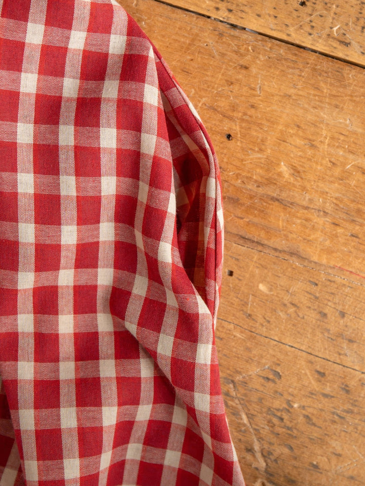 A red and white gingham cloth, inspired by the Savannah Sevens western life{&}style Tolland Red Gingham Button Up Dress, is draped over a wooden surface with visible grain and small marks.