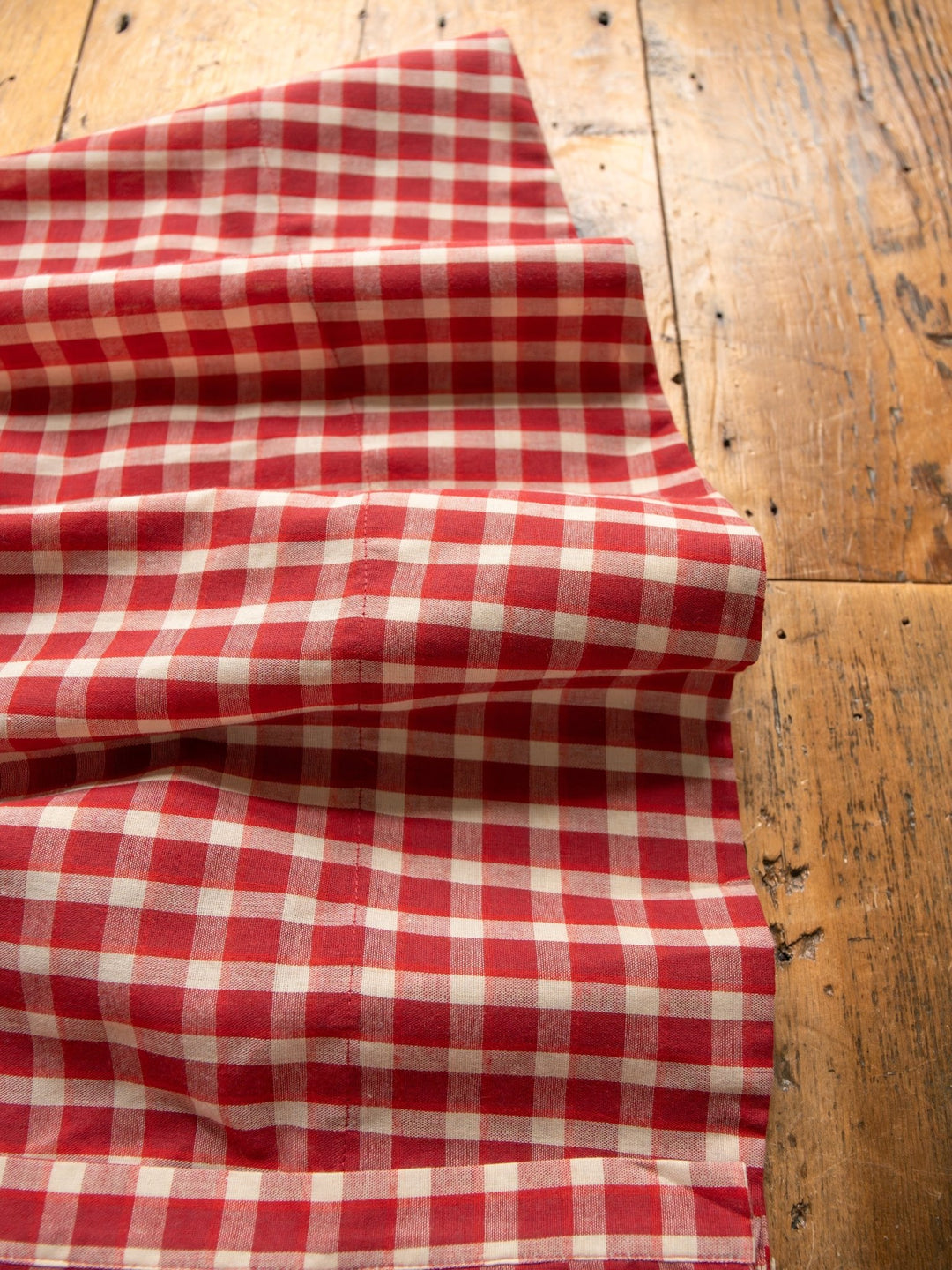 The Tolland Red Gingham Button Up Dress by Savannah Sevens western life{&}style, featuring a classic red and white checkered grid, is draped over rustic wood with visible grain and knots.