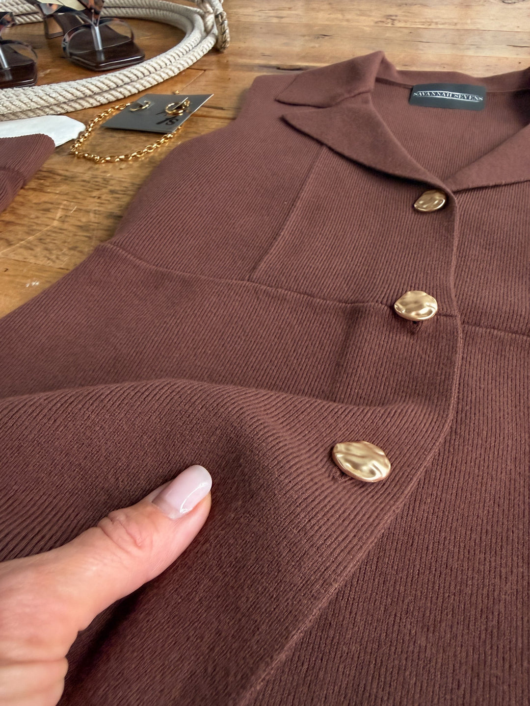 A person holds the ribbed tri-blend fabric of the Tolleson Chocolate Knit Vest Set by Savannah Sevens western life&style, highlighting its texture and detail. Accessories, such as a gold chain and sunglasses, are partially visible on a wooden table.