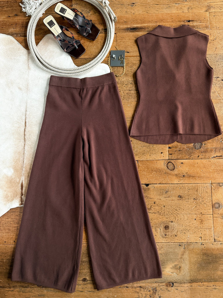 The Tolleson Chocolate Knit Vest Set by Savannah Sevens western life{&}style, featuring a vest top and wide-leg pants, is laid flat on a wooden floor alongside brown sandals, a white rope, and a cowhide rug.