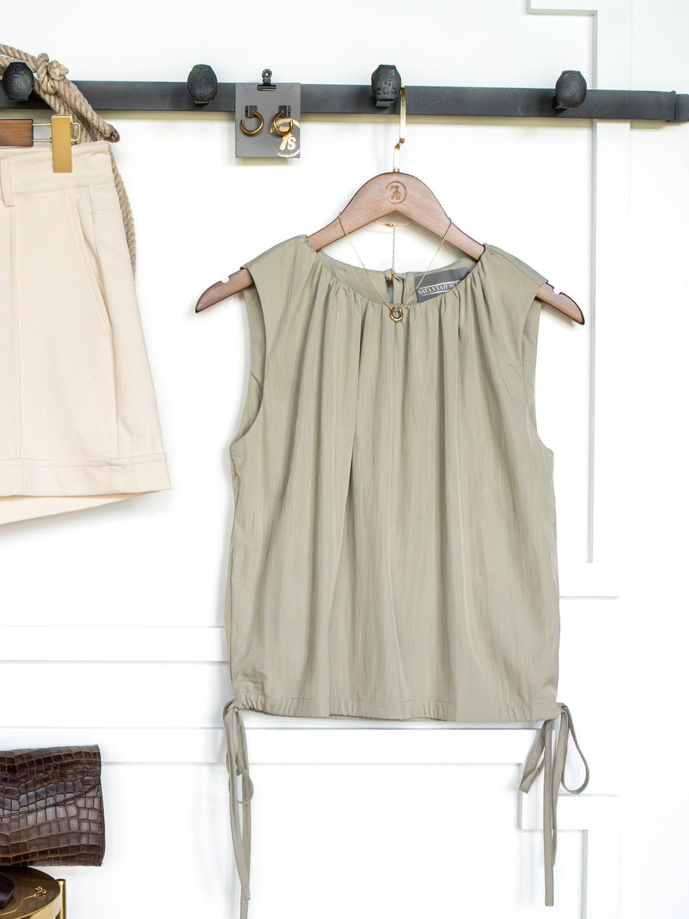 The Tolley Sage Side Tie Top by Savannah Sevens western life{&}style, featuring a ruched neckline and adjustable side tie, hangs on a wooden hanger against a white wall. Beige shorts, a brown bag, and rope belt are partially visible nearby.