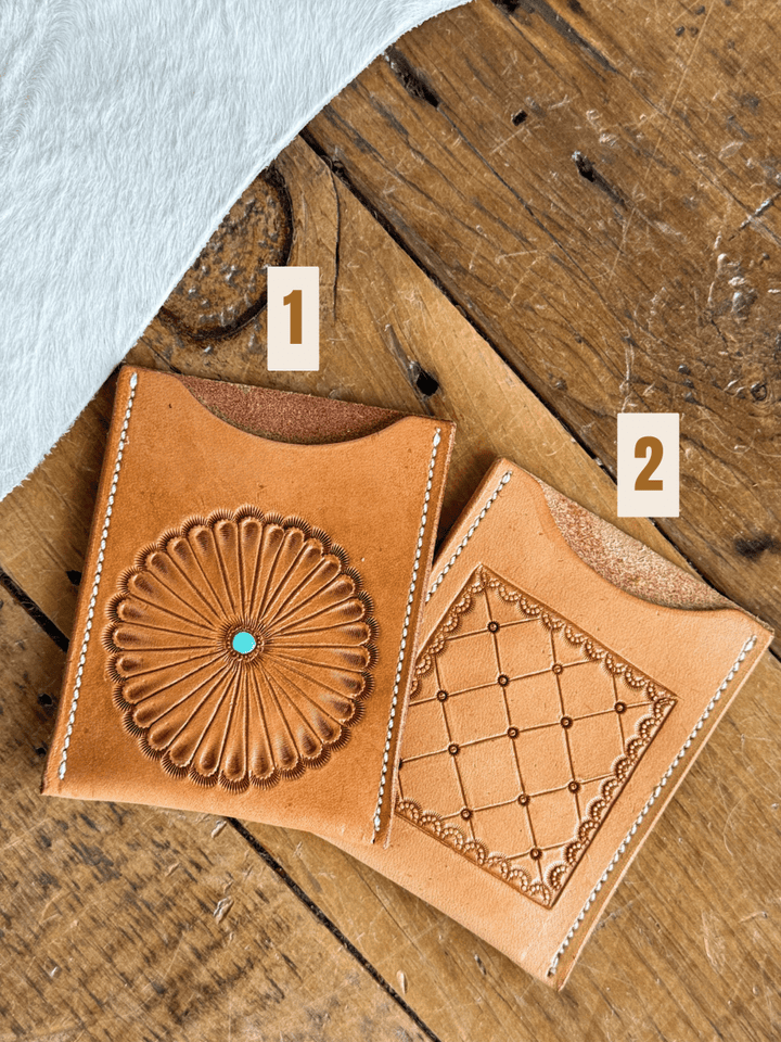 Two Tooled Card Wallets by Savannah Sevens western life{&}style rest on wood: Wallet 1 shows a sunburst with turquoise center, Wallet 2 displays a diamond pattern. Both feature white stitching and are labeled 1 and 2.