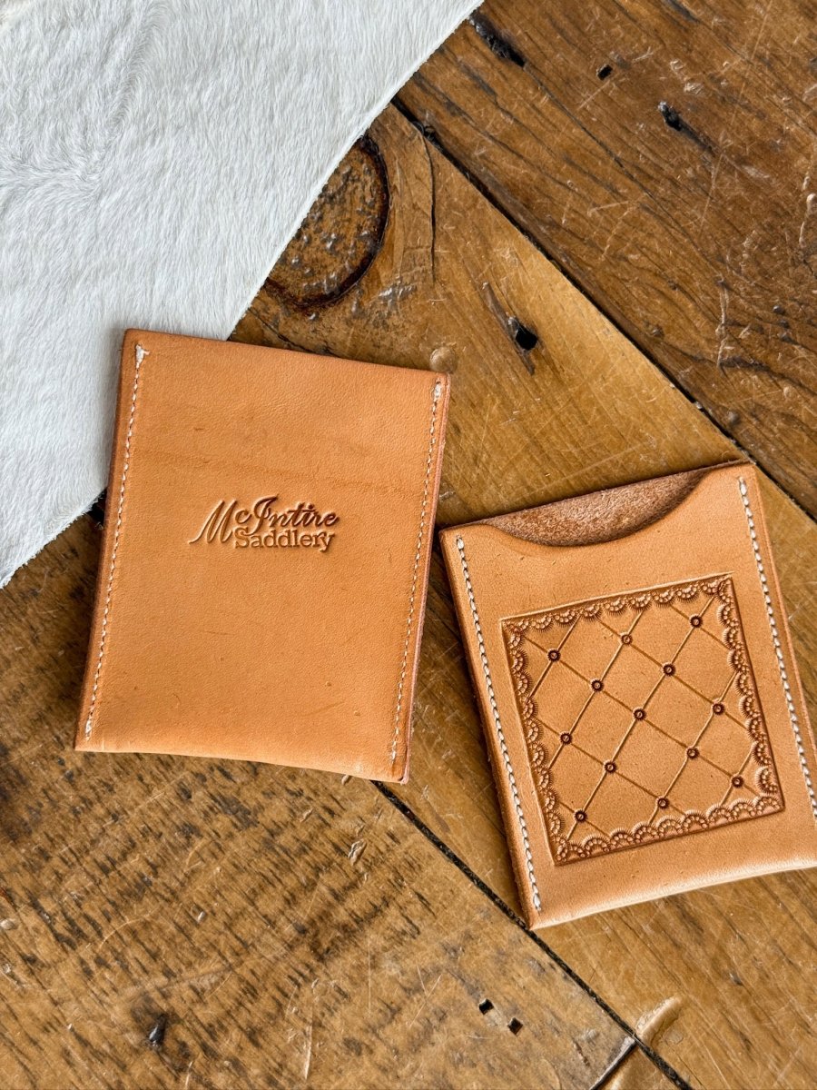 Two tan leather Tooled Card Wallets from Savannah Sevens western life{&}style rest on wood; one is embossed with McIntire Saddlery, the other shows a stitched geometric pattern with dot details. A white fur piece is partially visible nearby.