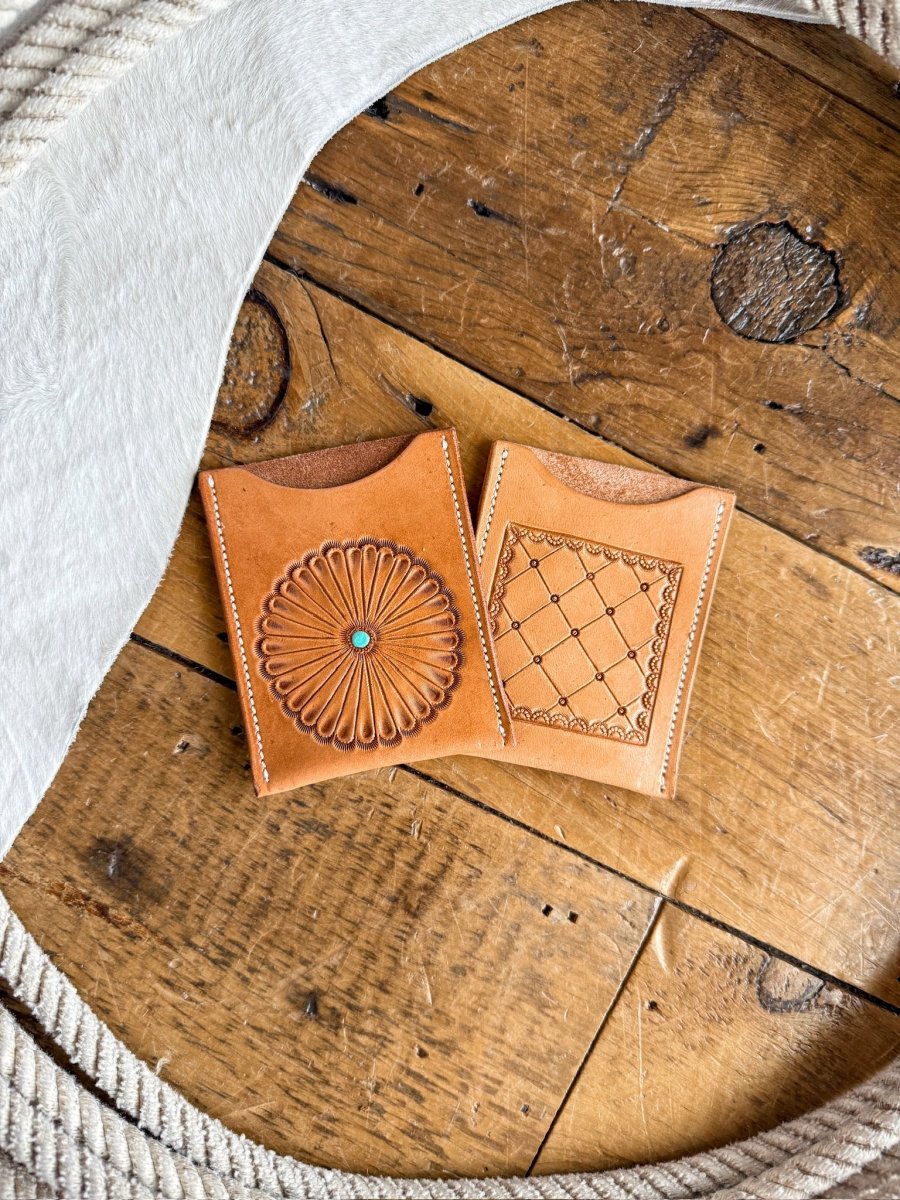 Two tan leather Tooled Card Wallets by Savannah Sevens western life{&}style sit on a wooden surface. One features a floral pattern with a turquoise center, the other has a diamond design. A rope and white rug appear in the corner.