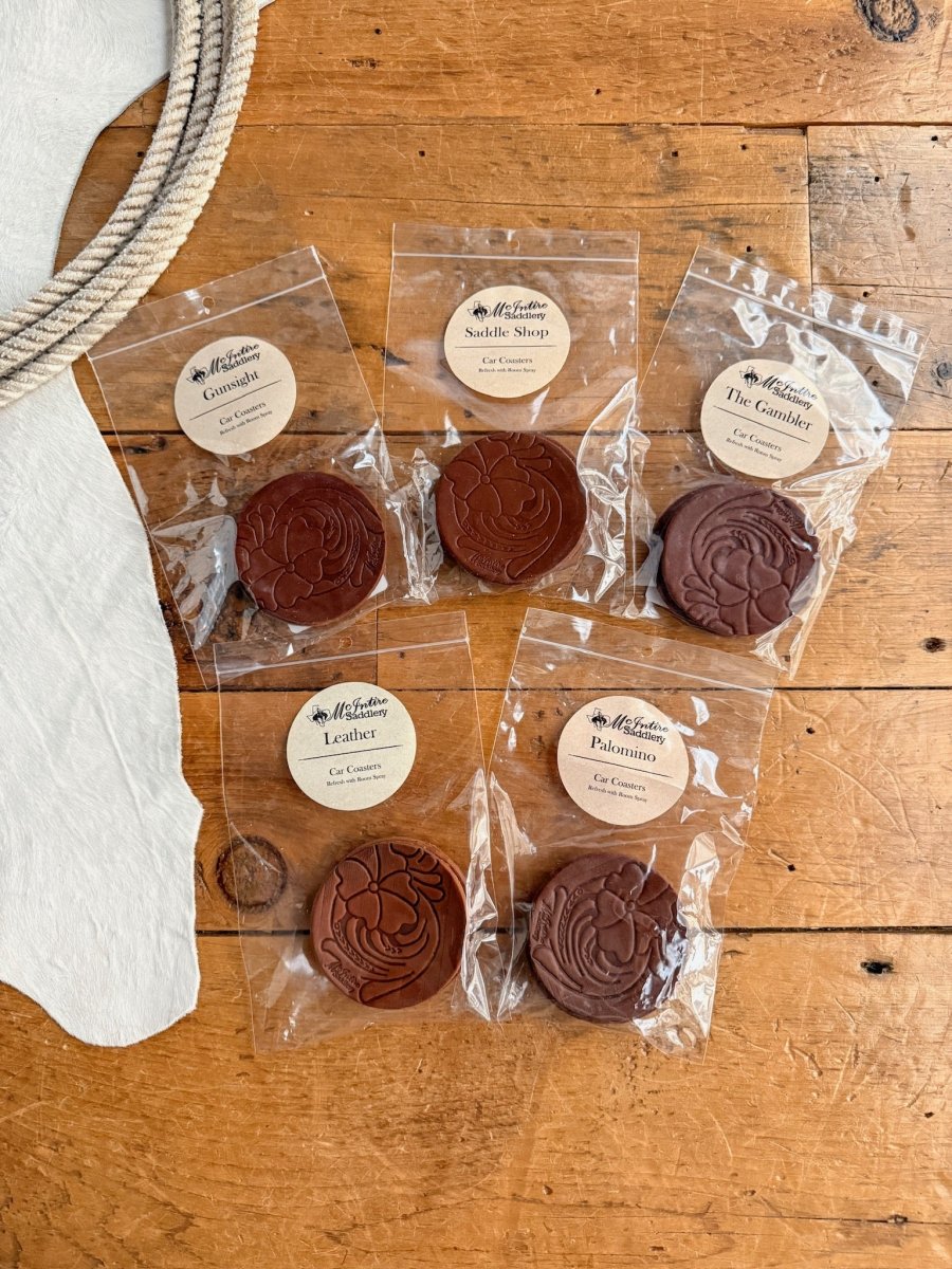 Six round Tooled Leather Car Coasters by Savannah Sevens western life{&}style, available in five scents like leather and Saddle Shop, are displayed in clear plastic packs with brown labels on a wooden surface beside rope and fabric.