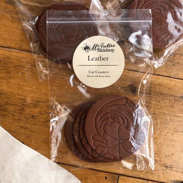 A clear plastic bag labeled "Tooled Leather Car Coaster {5 scents}" by Savannah Sevens western life{&}style contains brown, round tooled leather car coasters with embossed patterns, displayed on a wooden surface.