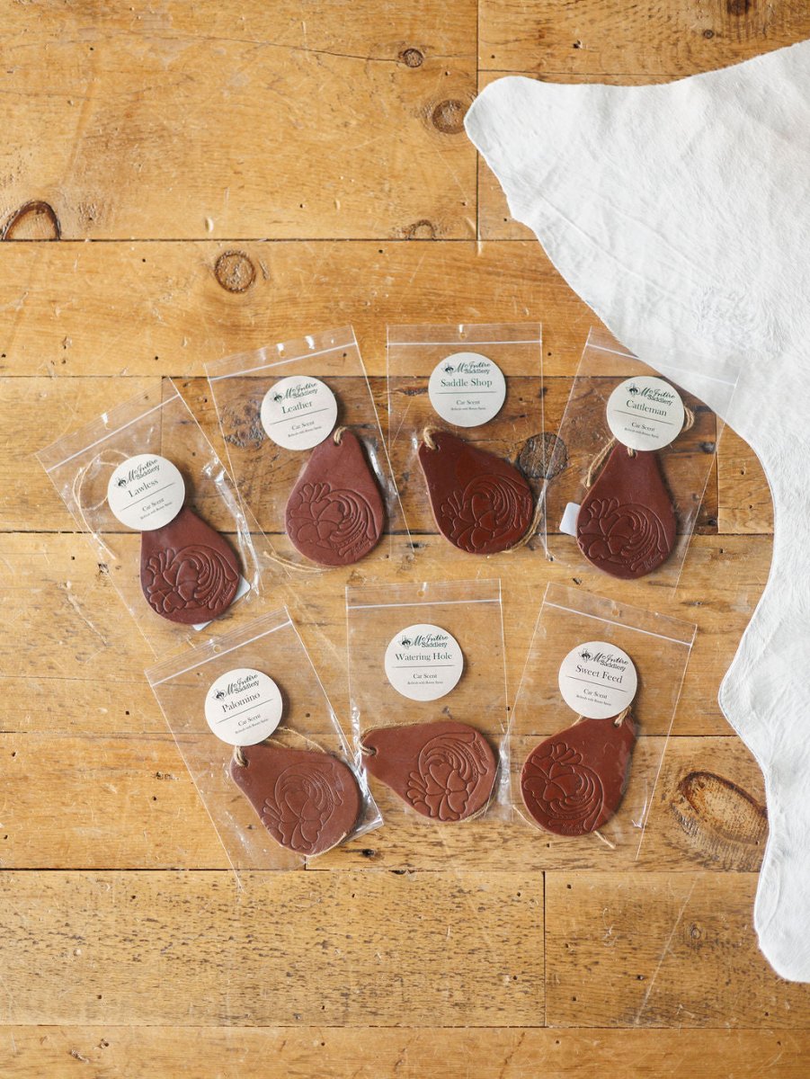 Seven Tooled Leather Car Fresheners by Savannah Sevens western life{&}style, each in teardrop shape with floral designs, are arranged on wood. Individually packaged in clear plastic with round labels. A white cloth is visible at top right.