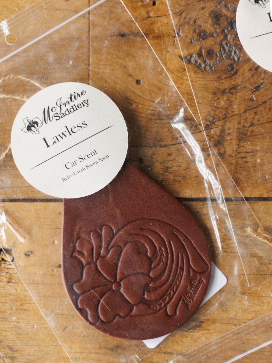 A Tooled Leather Car Freshener by Savannah Sevens western life{&}style, available in 7 scents, features a brown floral-embossed design and comes in clear packaging, displayed on a wooden surface.