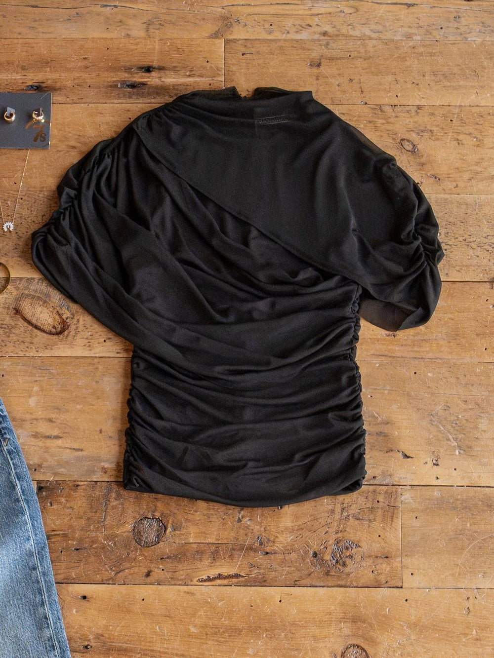 The Toxey Black Caplet Ruched Top by Savannah Sevens western life{&}style, featuring a sheer black fabric, asymmetrical design, and single sleeve, is laid flat on a wooden surface beside blue jeans and jewelry.