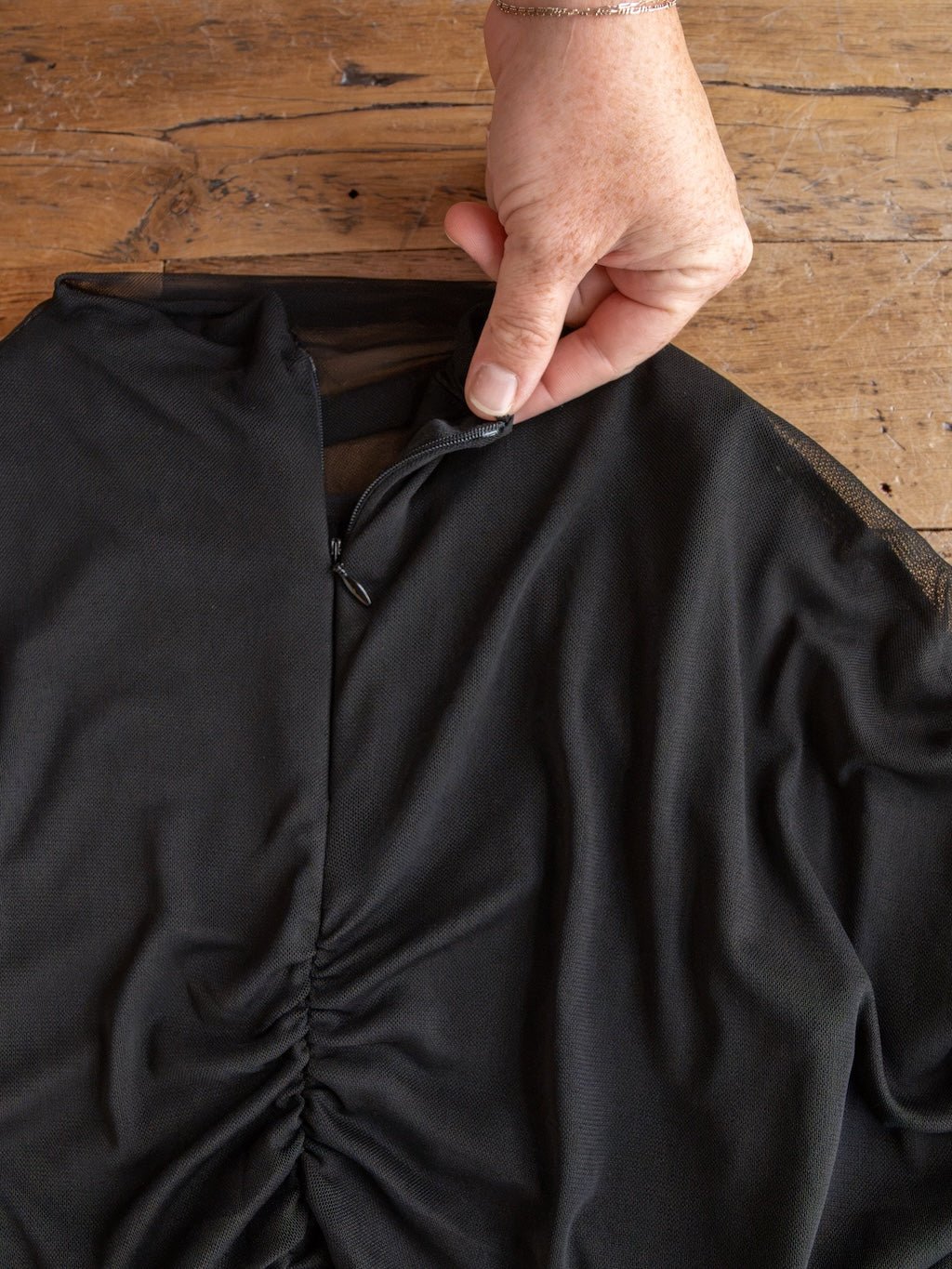 A hand holds the small black zipper on a gathered section of the Toxey Black Caplet Ruched Top by Savannah Sevens western life{&}style, placed on a wooden surface.