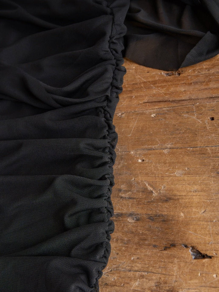 A close-up of the Toxey Black Caplet Ruched Top by Savannah Sevens western life{&}style, showcasing gathered black fabric and visible stitching, draped elegantly over a worn wooden surface.