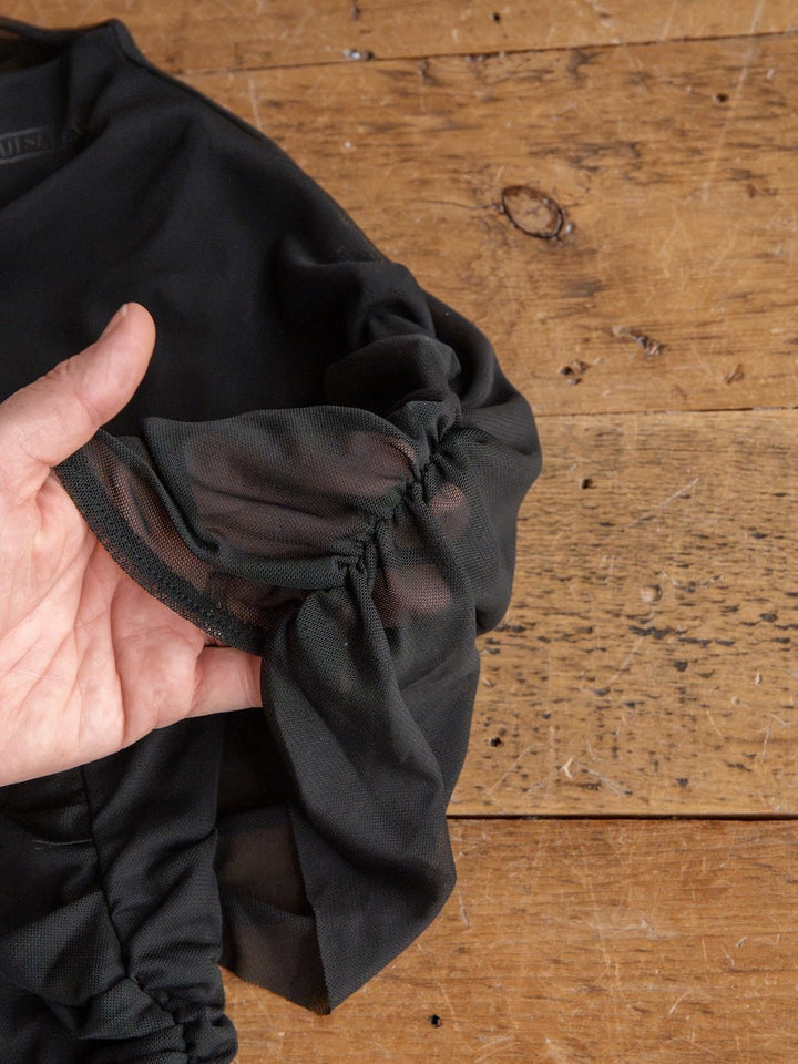 A hand holds the ruffled, sheer black sleeve of the Toxey Black Caplet Ruched Top by Savannah Sevens western life{&}style, displayed against a rustic wooden surface.