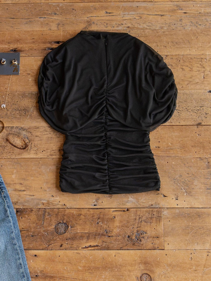The Toxey Black Caplet Ruched Top by Savannah Sevens western life{&}style is laid flat on a wooden surface, next to the corner of a pair of blue jeans and a jewelry tray.