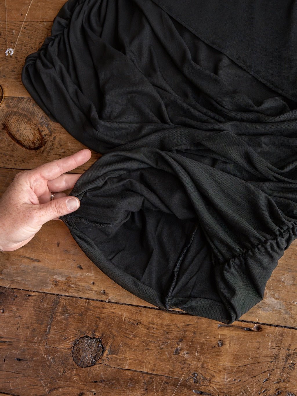 A hand gently holds the gathered edge of the Savannah Sevens western life{&}style Toxey Black Caplet Ruched Top on a rustic wooden surface, with jewelry partially visible in the corner.