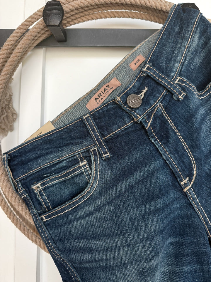 A pair of Trego Perfect Rise Trouser Jeans by Savannah Sevens western life{&}style, featuring a medium dark wash with tan stitching, hangs on a rope hanger against a white background.