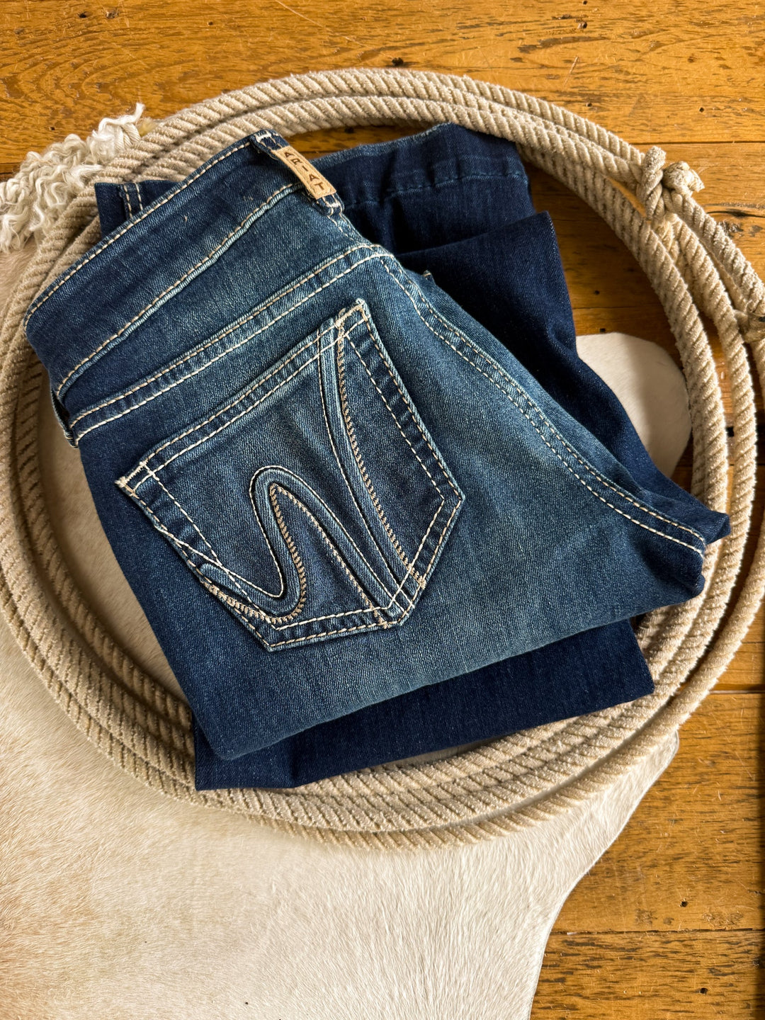 Two pairs of folded Savannah Sevens western life{&}style Trego Perfect Rise Trouser Jeans in a medium dark wash sit atop coiled rope and a white cowhide rug, all placed on a wooden floor.