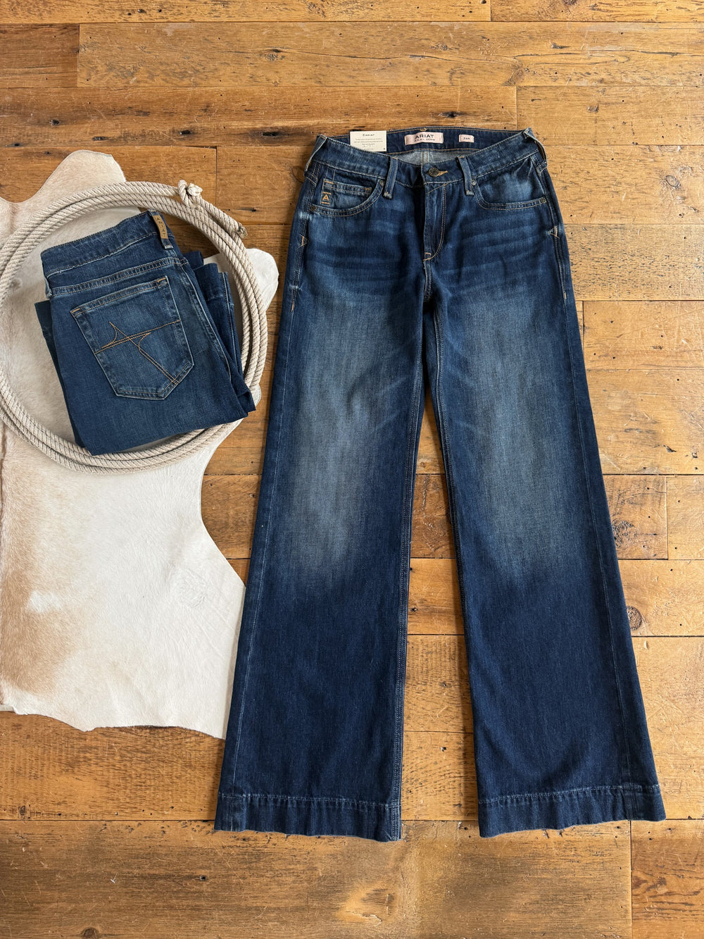 Tryan Perfect Rise Wide Leg Jeans by Savannah Sevens western life{&}style in a medium dark wash are laid flat on a wooden floor next to a folded pair of jeans, a coiled rope, and a cowhide rug.