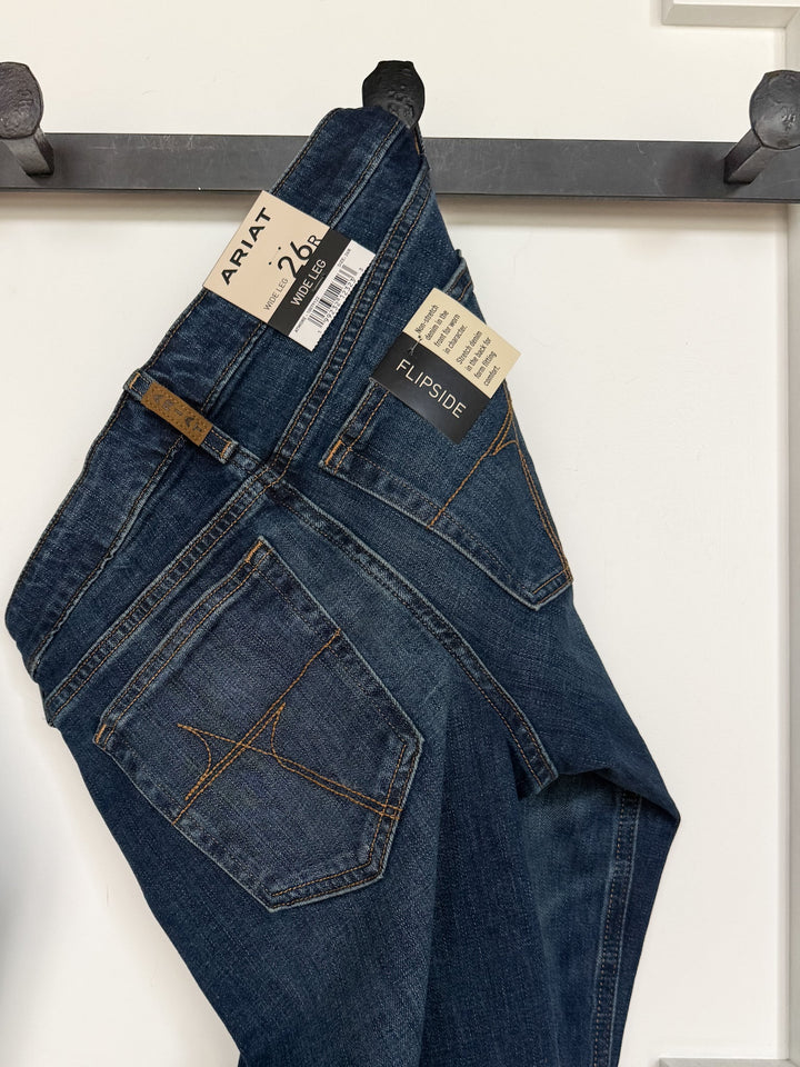 A pair of blue Tryan Perfect Rise Wide Leg Jeans from Savannah Sevens western life{&}style hangs by a hook, displaying the back pockets, leather brand patch, and tags. These wide leg jeans feature a medium dark wash with orange stitching and showcase perfect rise denim for a flattering fit.